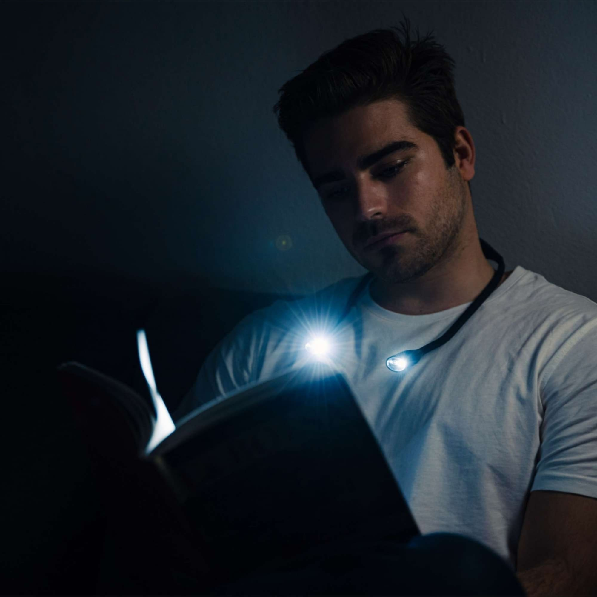 Hombre leyendo libro con luz de cuello en habitación oscura. Luz en libro y cara del hombre.