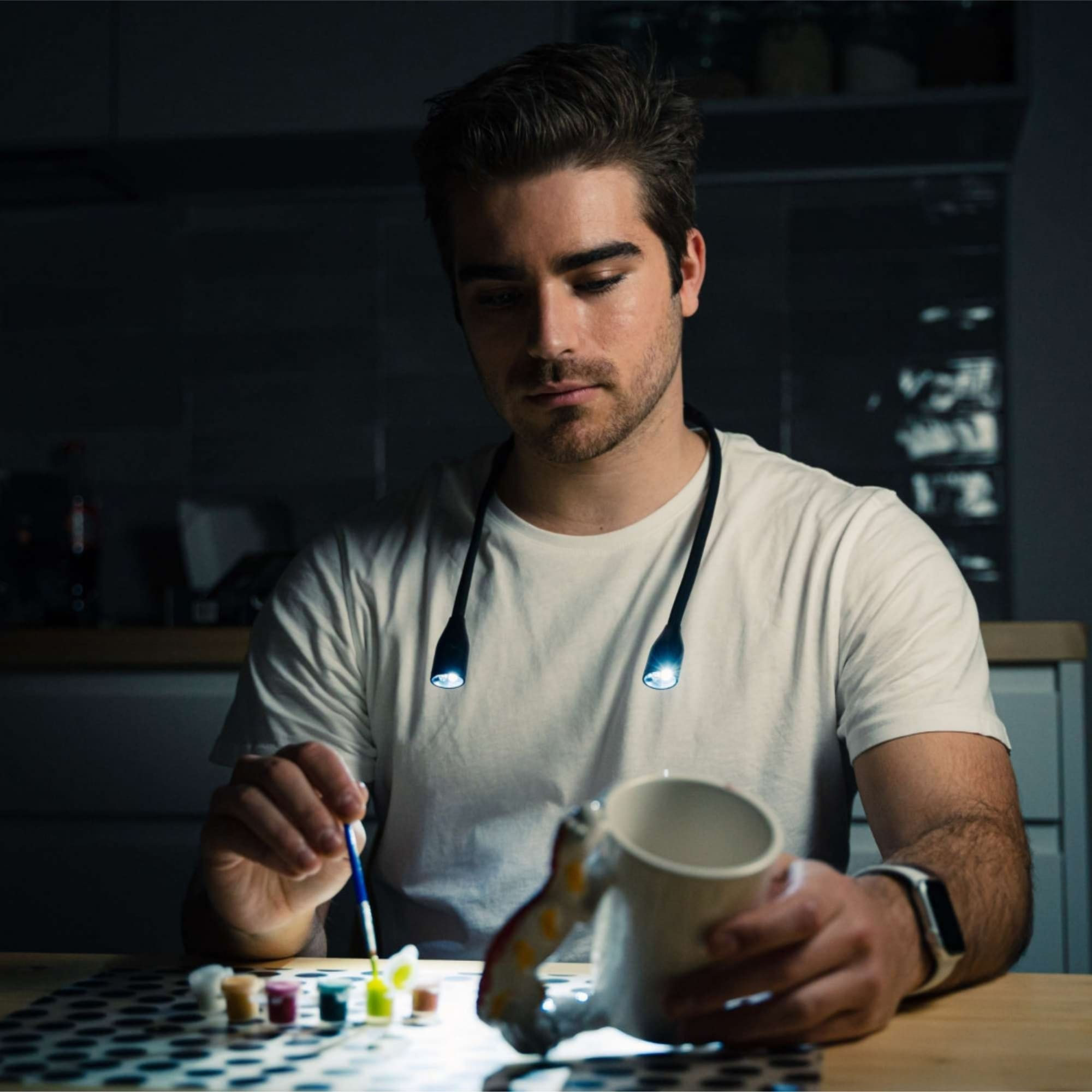 Hombre pintando con una luz de cuello. Está en una habitación con una taza blanca y pintura.