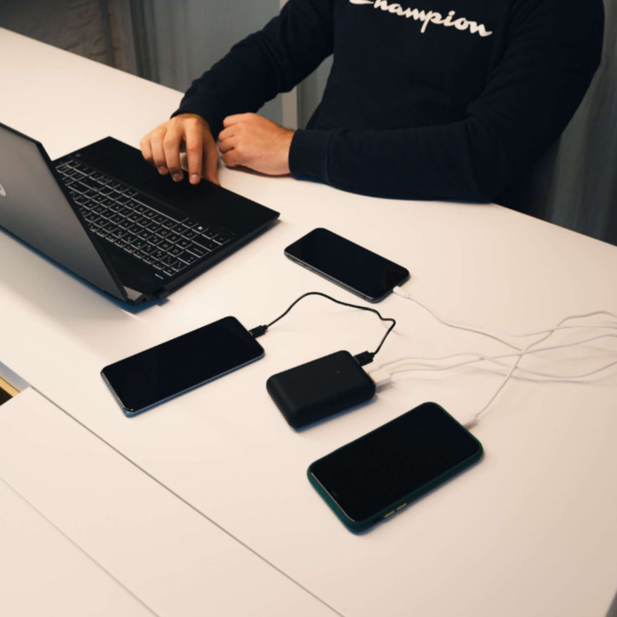 Una persona usando una laptop. Hay tres teléfonos y un banco de energía en la mesa. La mesa es blanca.