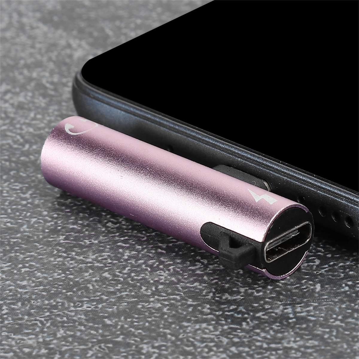 Ein rosa USB-C-Adapter, der an ein schwarzes Telefon angeschlossen ist, auf einer grauen Oberfläche. Der Adapter hat ein Logo.