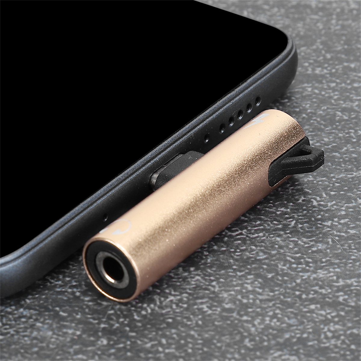 Ein goldfarbener Adapter ist in ein schwarzes Telefon eingesteckt. Es hat eine Kopfhörerbuchse. Das Telefon liegt auf einer strukturierten Oberfläche.