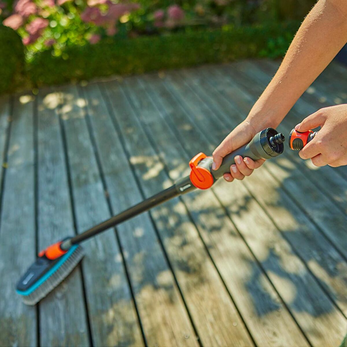 Een persoon verbindt een zwart-oranje tuinborstel met een waterslang op een houten dek.