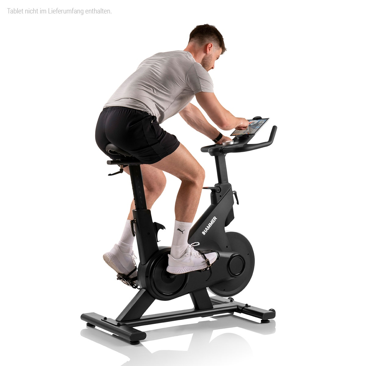 Homme en short et t-shirt sur un vélo d'exercice noir, utilisant une tablette montée sur le guidon.