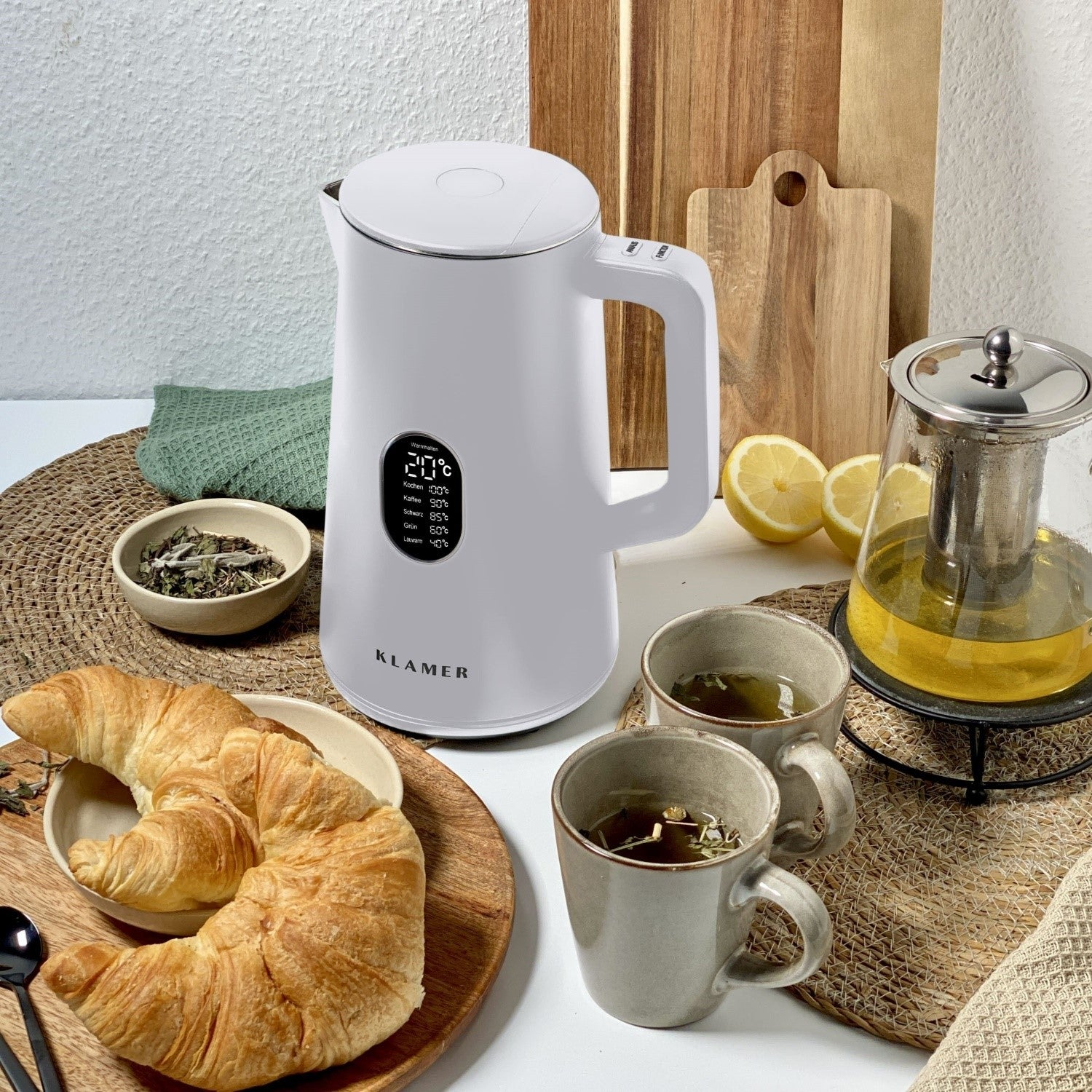 Weißer Wasserkocher mit Digitalanzeige, Croissants, Tee und Tassen auf dem Tisch.