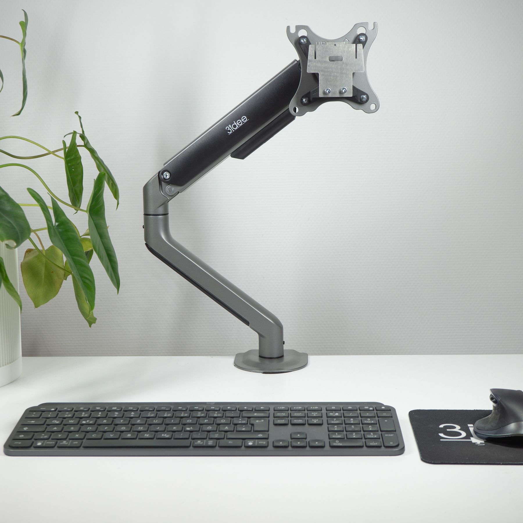 Grauer Monitorarm mit Monitor, Tastatur, Maus und Pflanze auf weißem Schreibtisch.