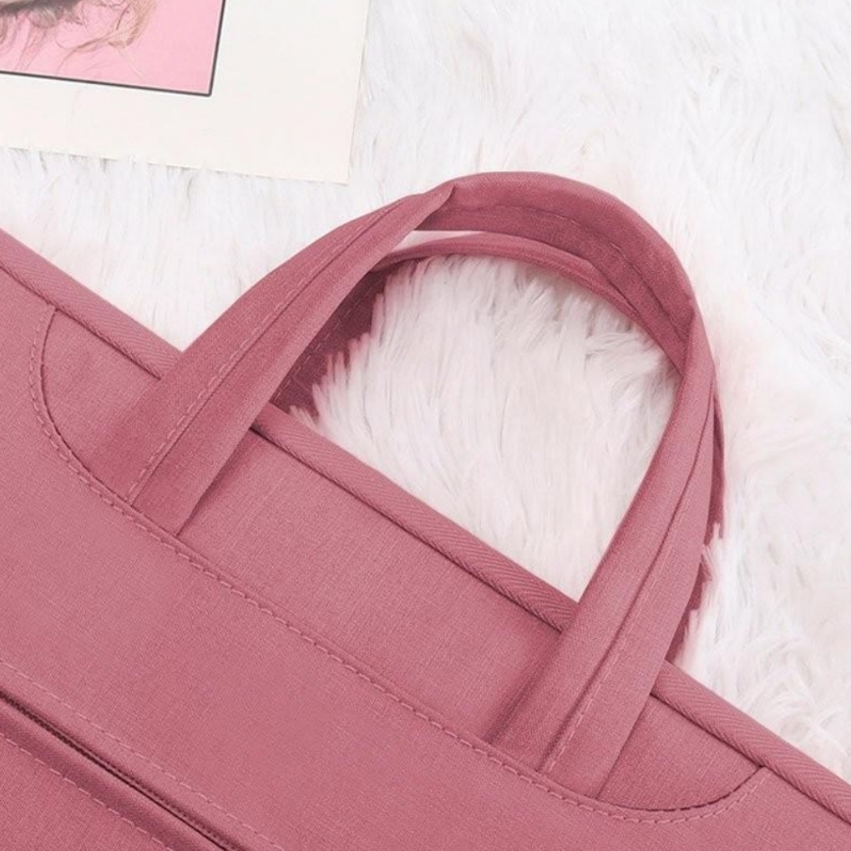 Rosafarbene Laptoptasche mit Griffen auf einer weißen, flauschigen Oberfläche. Ein Bild im Hintergrund.