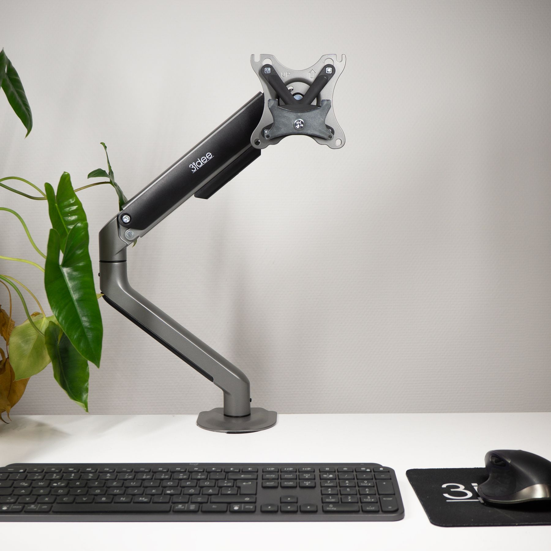 Monitorarm auf einem Schreibtisch, mit Tastatur, Maus und Pflanze im Hintergrund.