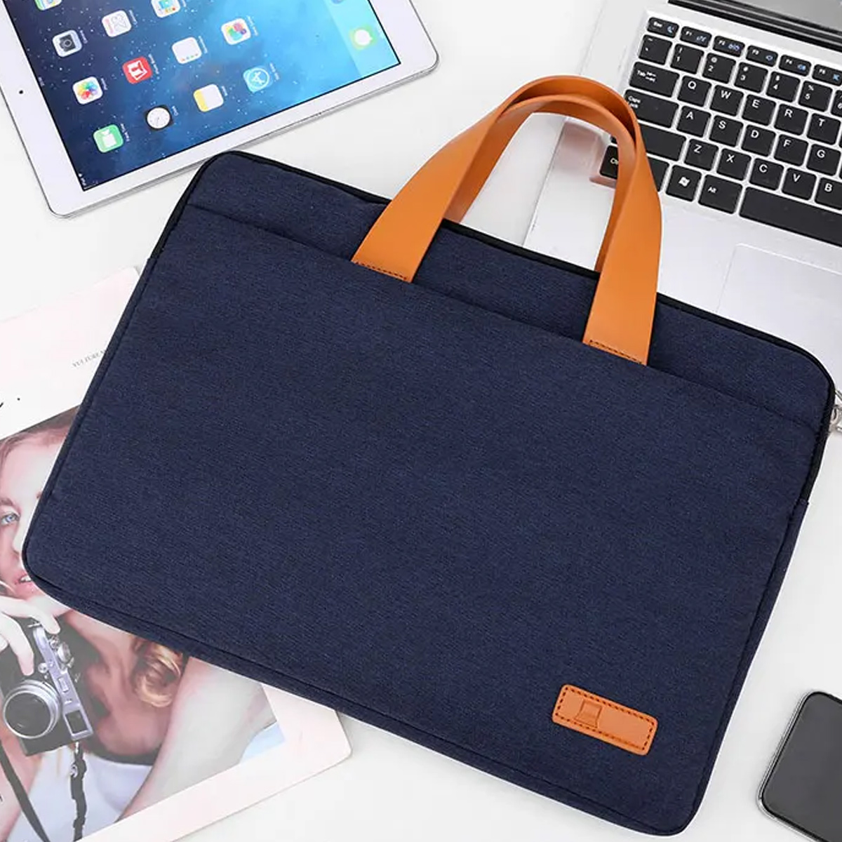 Eine blaue Laptoptasche mit braunen Griffen, auf einer weißen Oberfläche mit Tablet und Laptop.