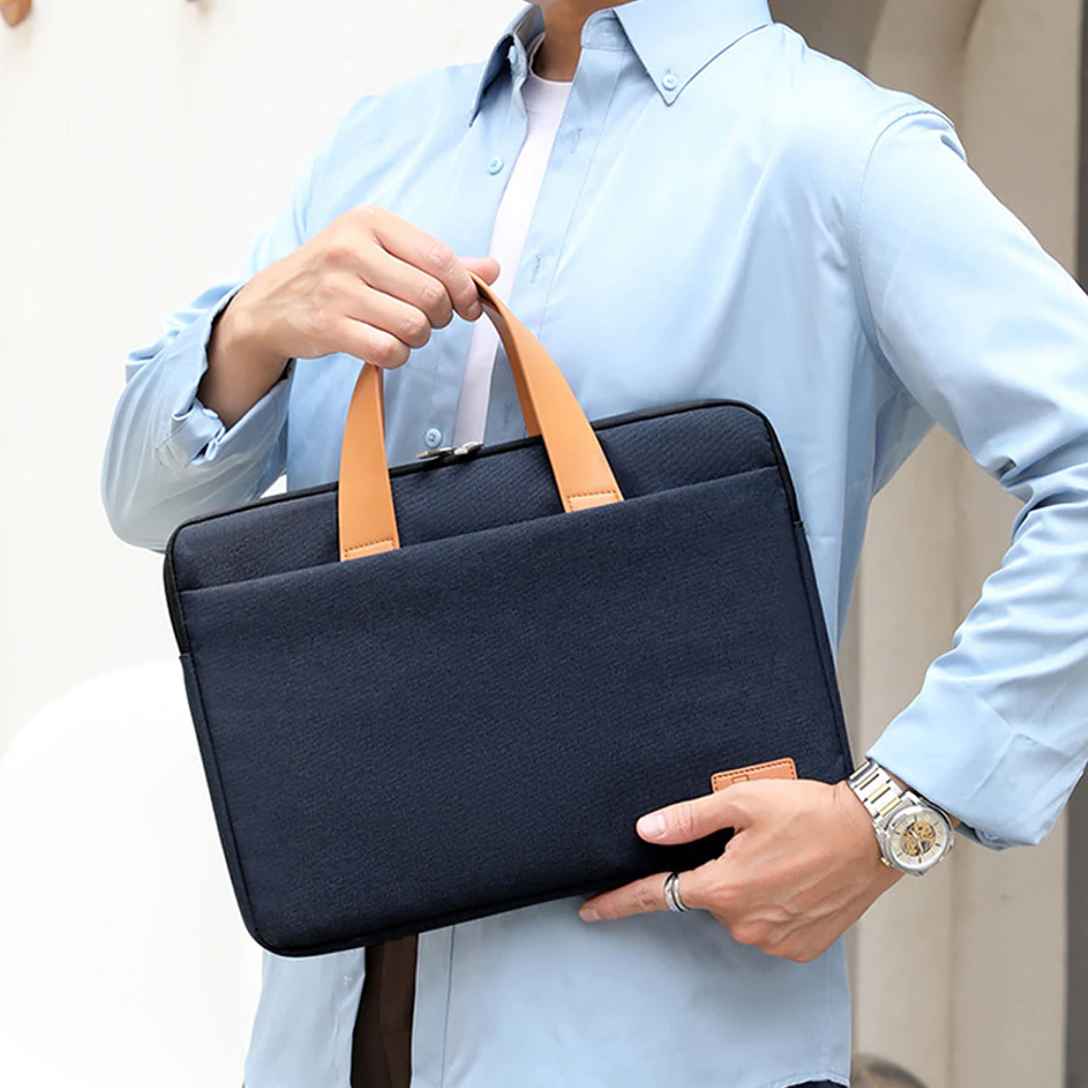 Eine Person hält eine marineblaue Laptoptasche. Die Tasche hat hellbraune Griffe und ein kleines Etikett. Person trägt ein blaues Hemd.