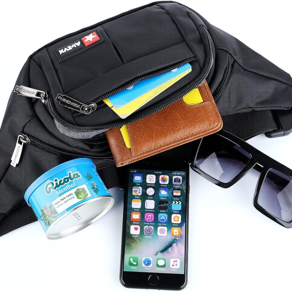 Schwarze Bauchtasche mit Gegenständen auf weißer Oberfläche. Zu den Gegenständen gehören ein Telefon, eine Sonnenbrille, eine Brieftasche und Süßigkeiten.