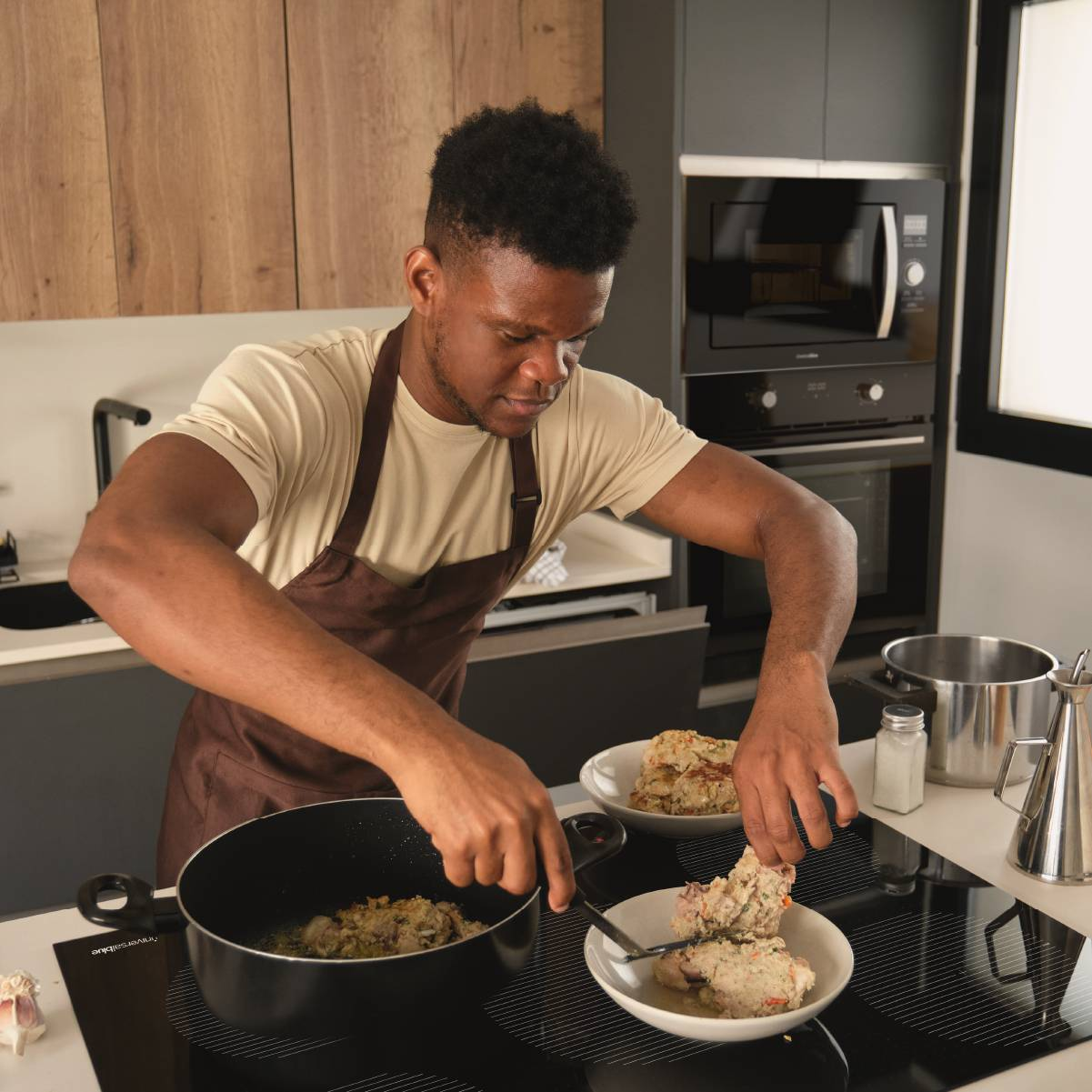 Hombre cocinando, colocando comida en un plato. Cocina con estufa, microondas e ingredientes.