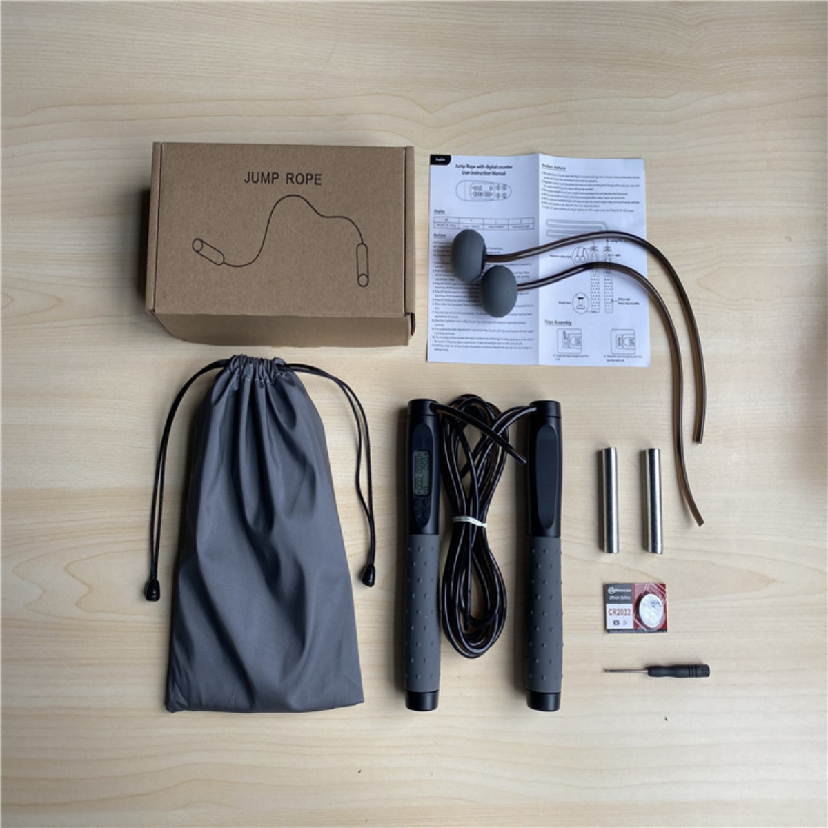 Springseil-Set mit Box, grauer Tasche, Springseil, Griffen, Gewichten und Batterie auf Holzoberfläche.