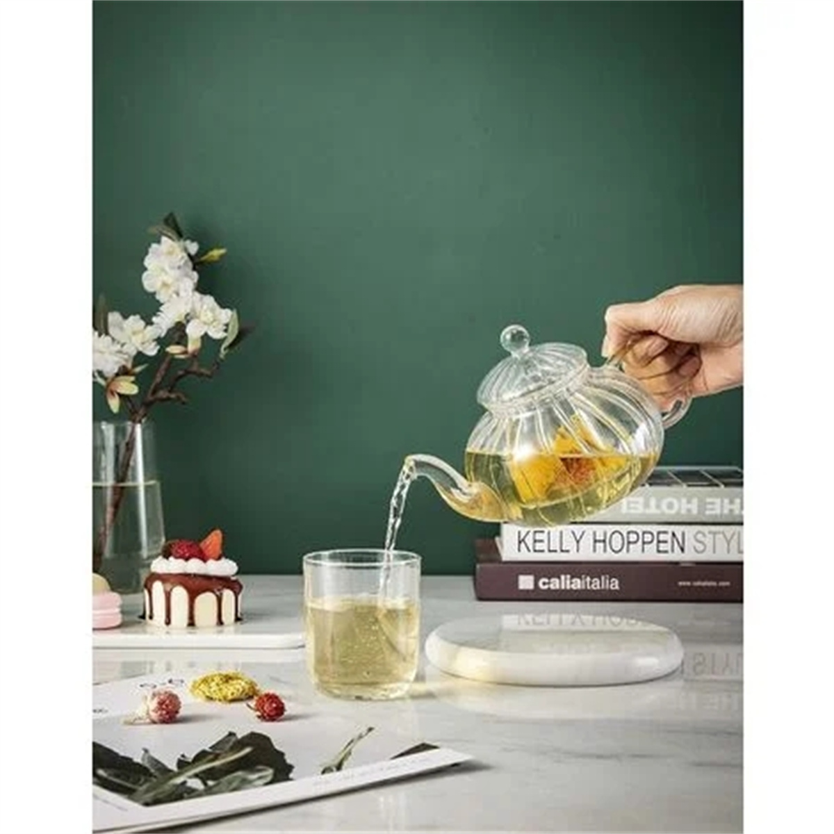 Eine Teekanne gießt Tee in ein Glas. Auf dem Tisch stehen Bücher, ein Kuchen und Blumen. Grüner Hintergrund.