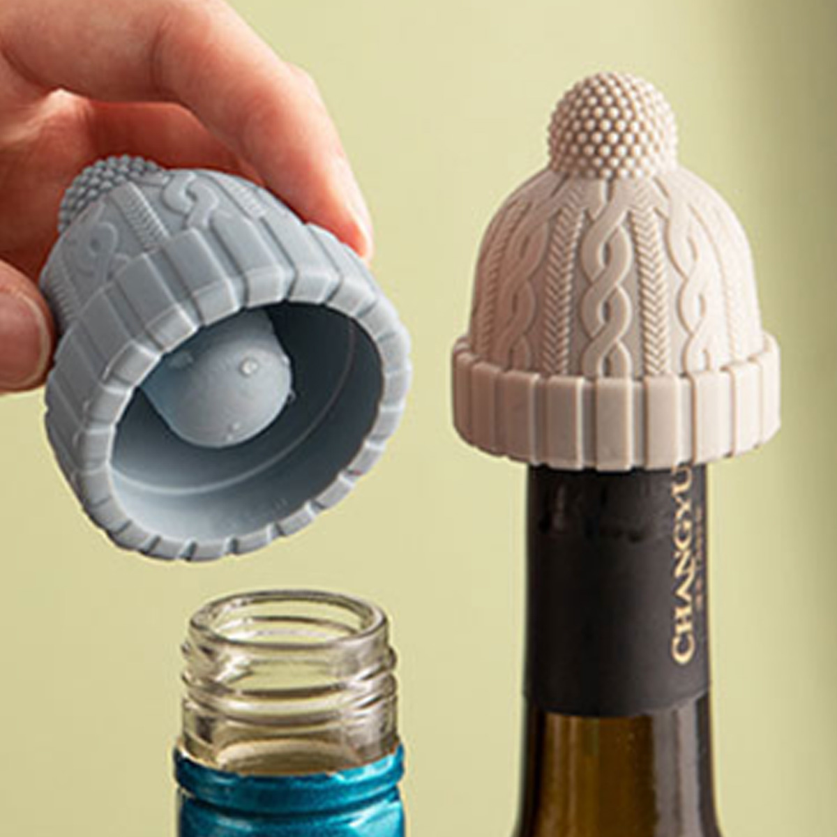 Zwei Weinflaschenverschlüsse in Form von Strickmützen, einer auf einer Flasche, grüner Hintergrund.