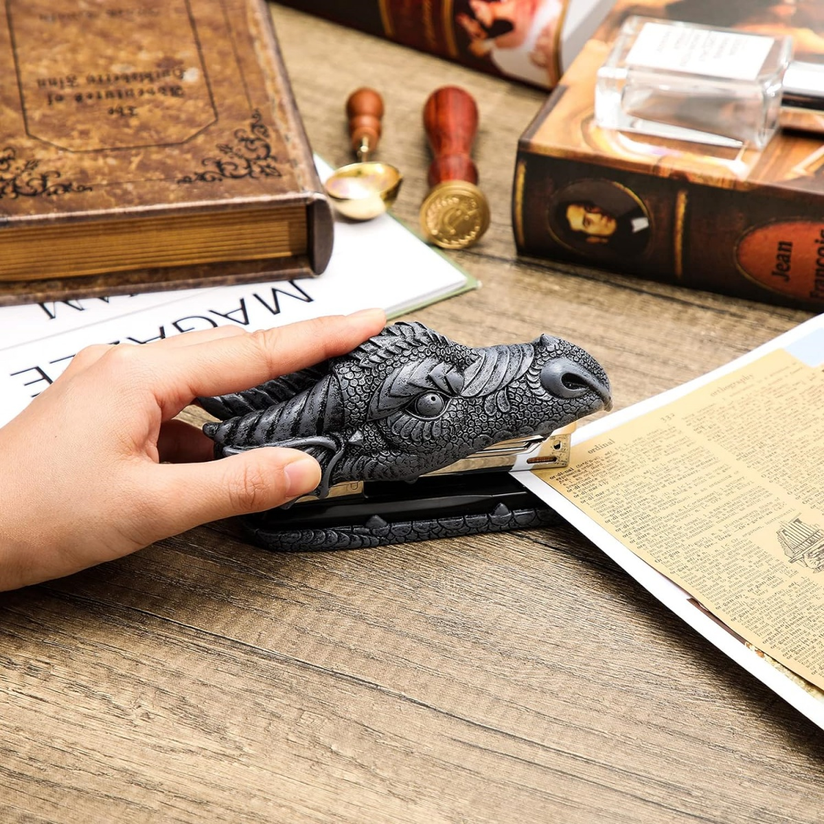 Die Hand einer Person benutzt einen drachenförmigen Tacker auf Papier. Weitere Gegenstände auf dem Tisch sind Bücher und Stempel.