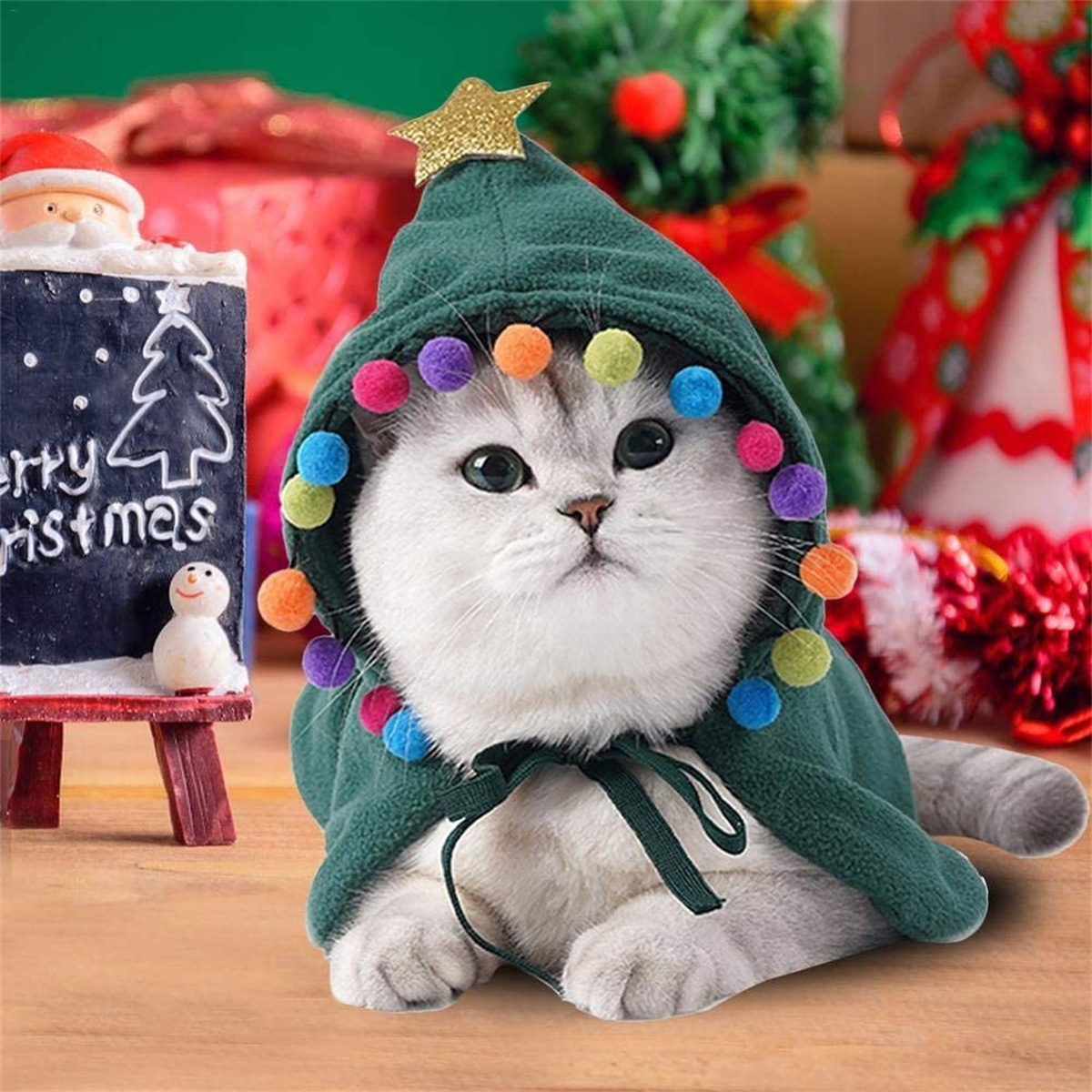 Katze mit grünem Weihnachtsbaumumhang und bunten Pompons, neben einer Tafel und Dekoration.