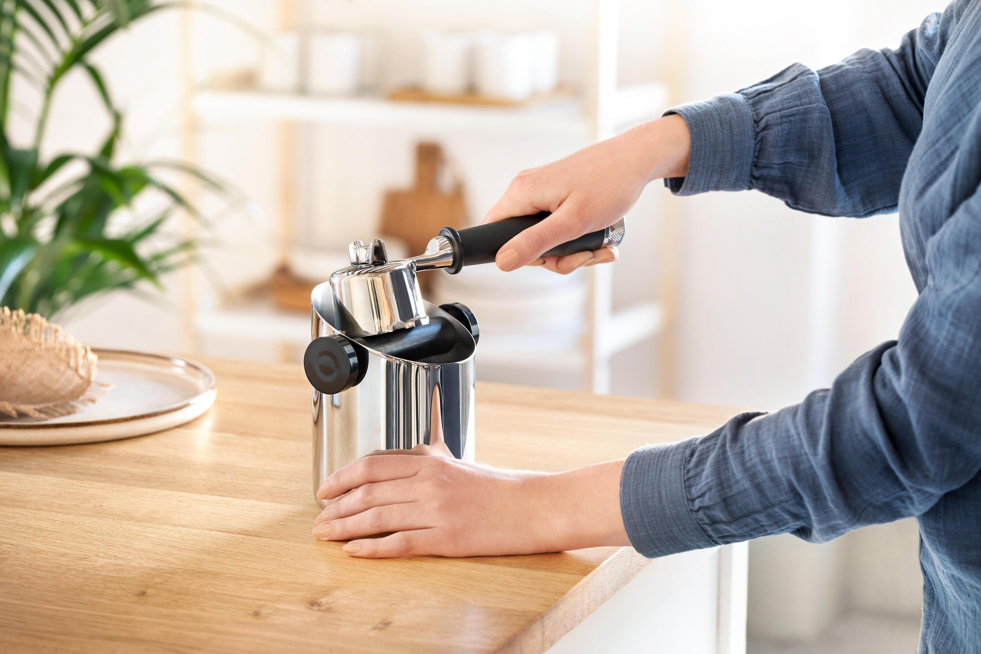 Hände halten eine silberne Kaffeemaschine über einem Holztisch, vor hellem Hintergrund.