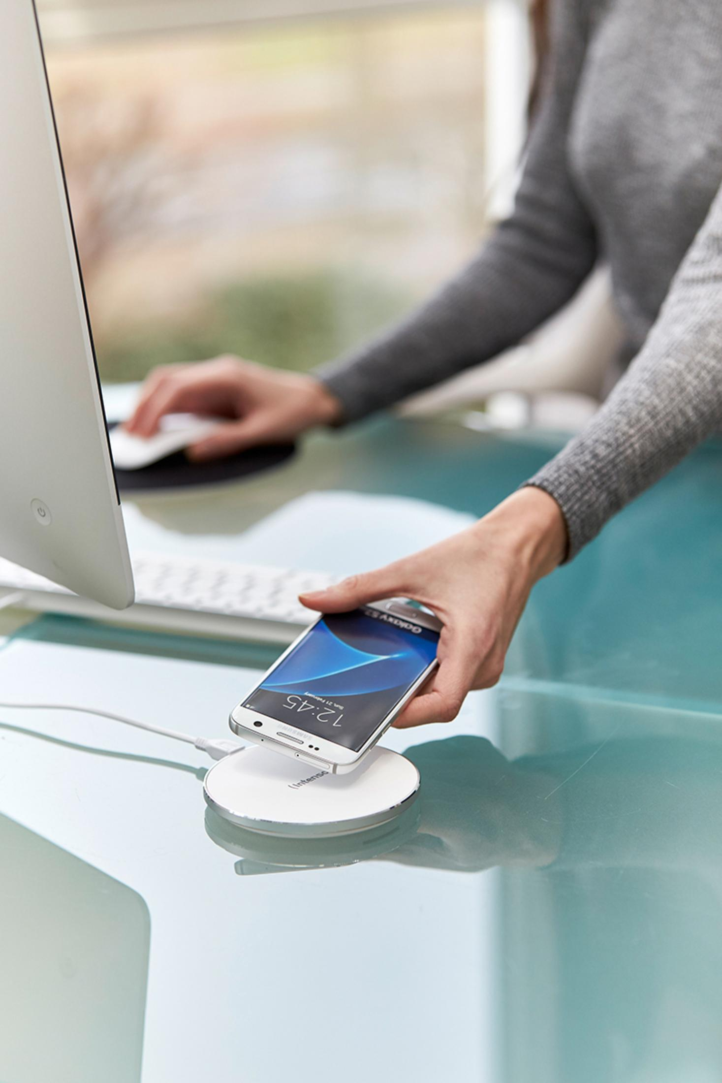 Hände einer Frau legen ein Telefon auf ein kabelloses Ladepad auf einem Glastisch in der Nähe eines Computers.