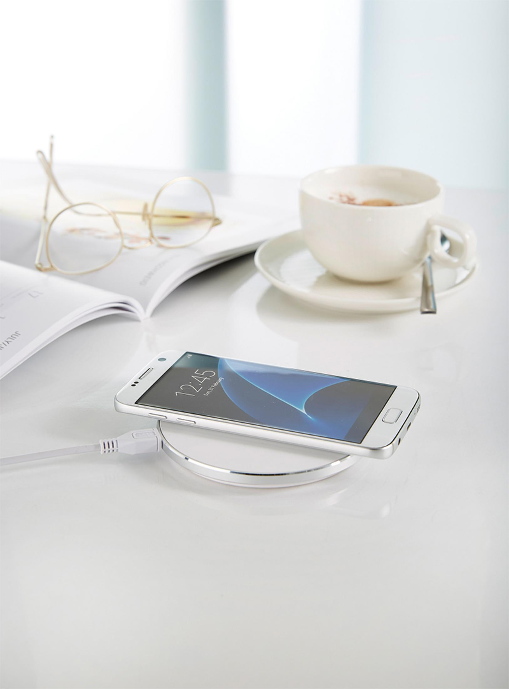 Ein weißes Telefon auf einem kabellosen Ladepad steht auf einem Tisch neben einer Tasse Kaffee und einer Brille.
