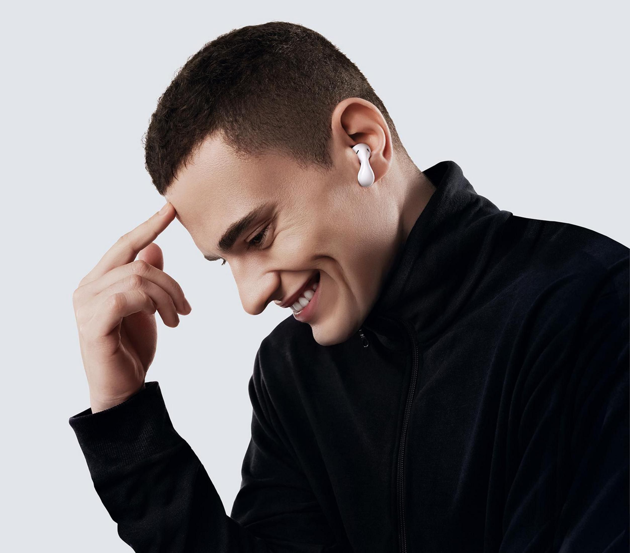 Mężczyzna w czarnej kurtce, uśmiechnięty i dotykający głowy, z białą słuchawką.