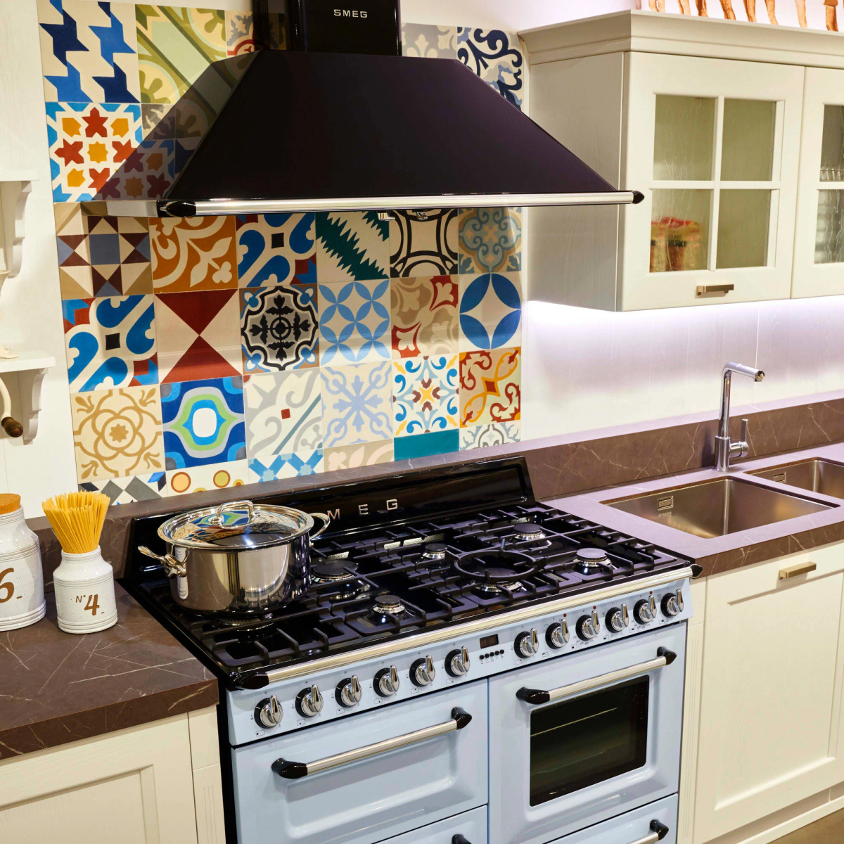 Una cucina con fornello blu, paraschizzi colorato e mobili bianchi, con una cappa nera.
