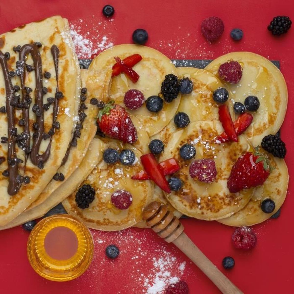 Naleśniki z syropem, jagodami i kawałkami czekolady na czerwonej powierzchni z miodem i nabierakiem do miodu.