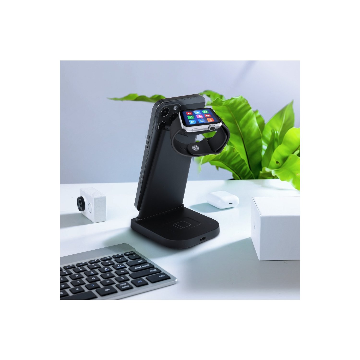 Schwarzer Ladeständer mit einer Apple Watch und einem Telefon, einer Tastatur und einer Pflanze im Hintergrund.