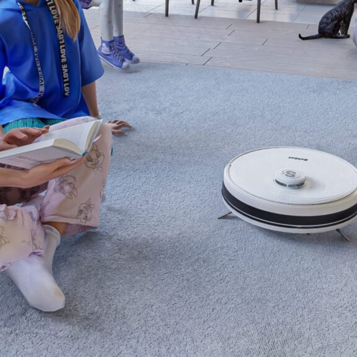 Eine Person liest ein Buch in der Nähe eines Roboter-Staubsaugers auf einem Teppichboden. Eine weitere Person sitzt daneben.