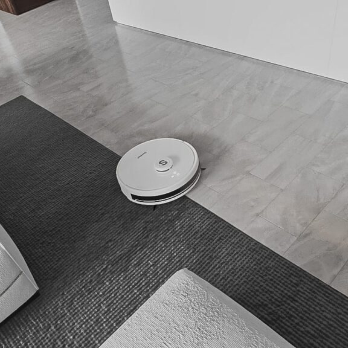 Ein weißer Roboter-Staubsauger auf einem Teppich und einem grauen Boden. Der Roboter ist nah an der Wand.