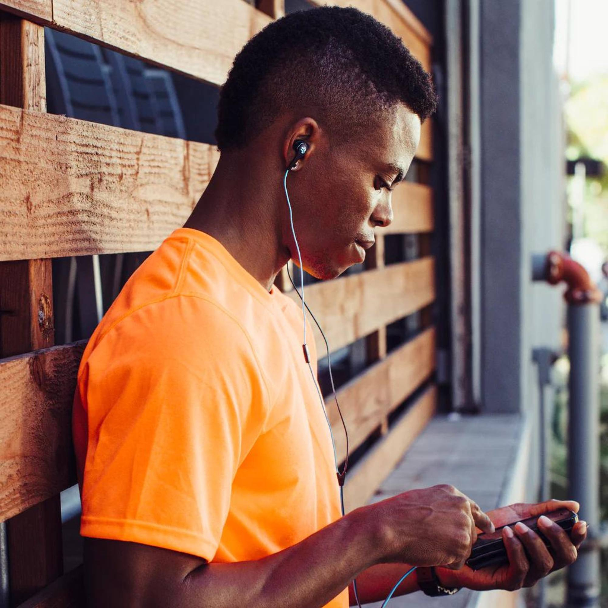 Joven con auriculares usando un teléfono, apoyado en una pared de madera.