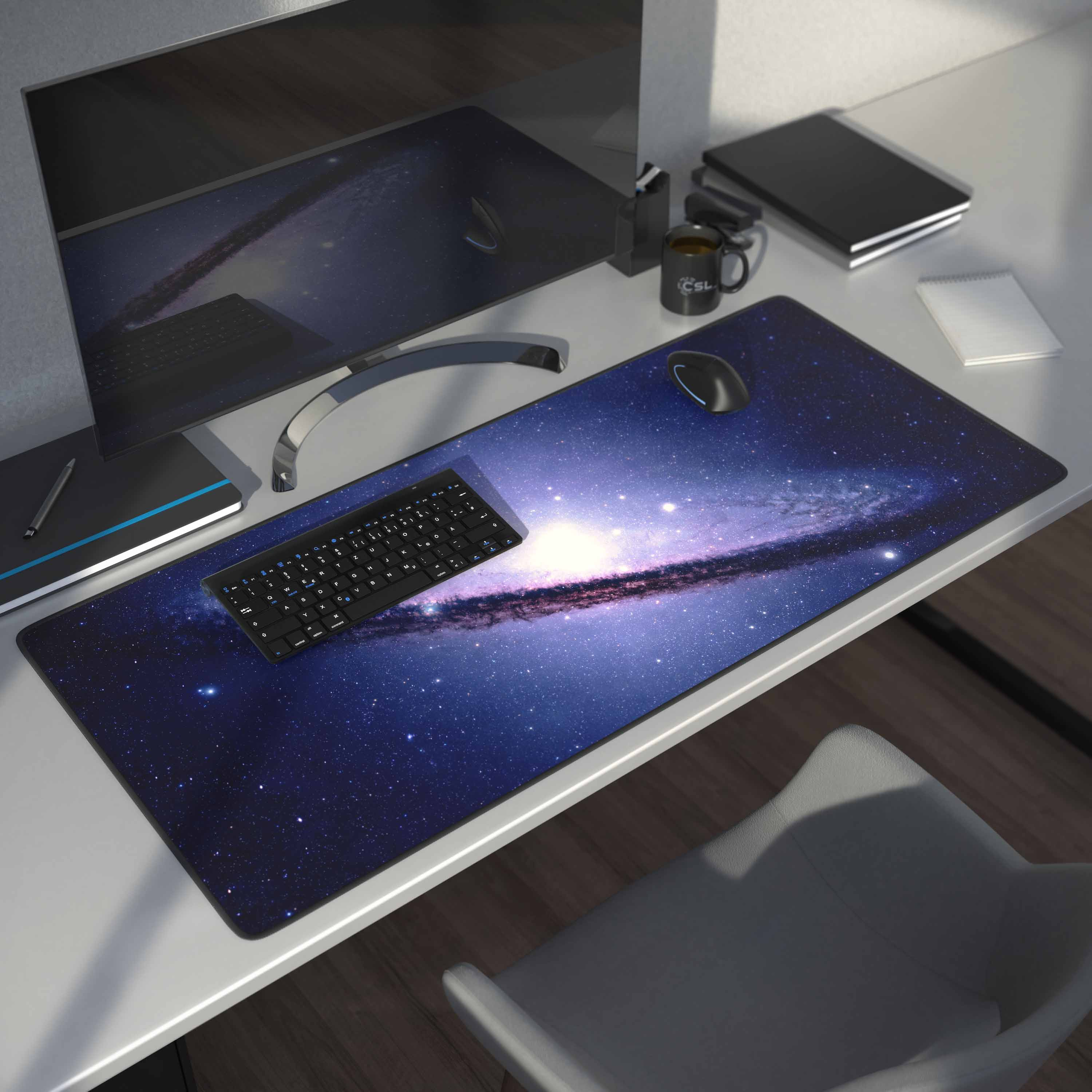 Ein Schreibtisch mit einem Mauspad im Galaxie-Design, Tastatur und Monitor, mit Tasse und Notizblock.