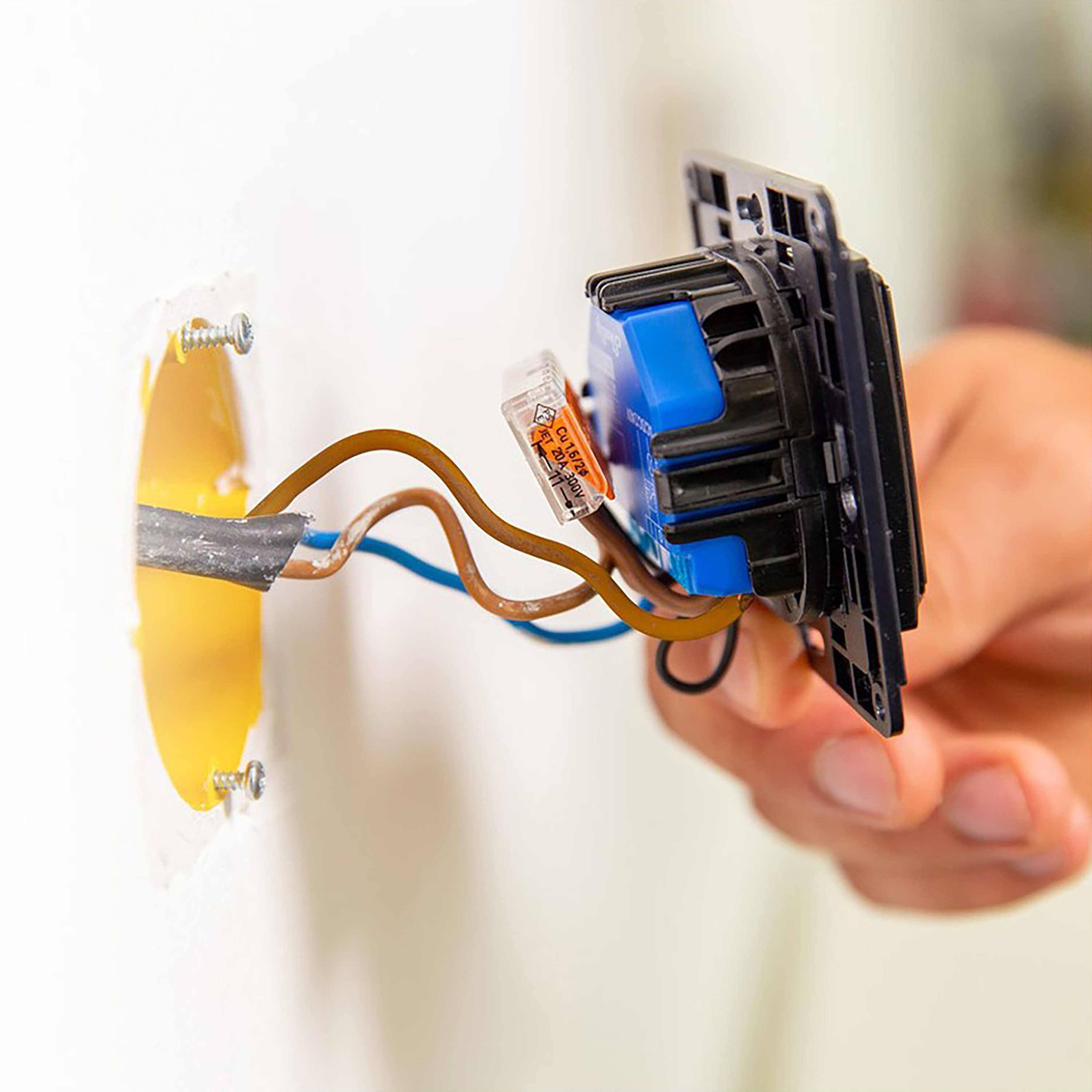 Una mano conecta cables a un interruptor. Los cables son marrones y azules. Pared blanca.