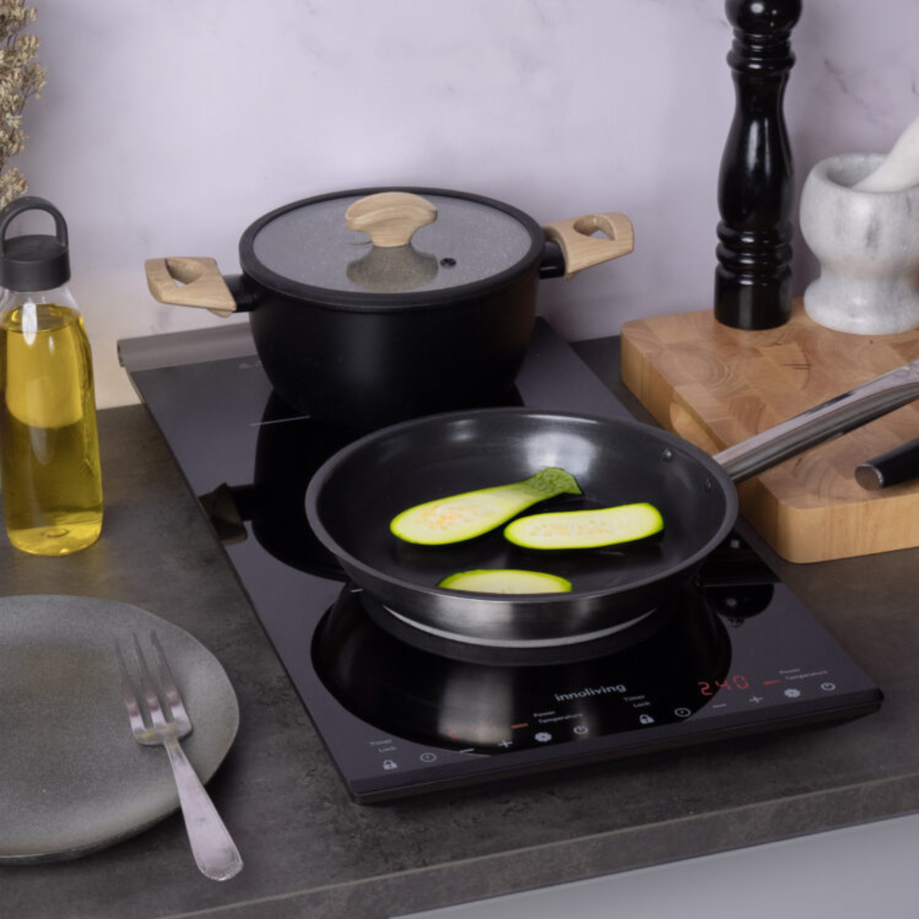 Configurazione di cottura con zucchine in una padella su un piano cottura a induzione, con una pentola e olio d'oliva.