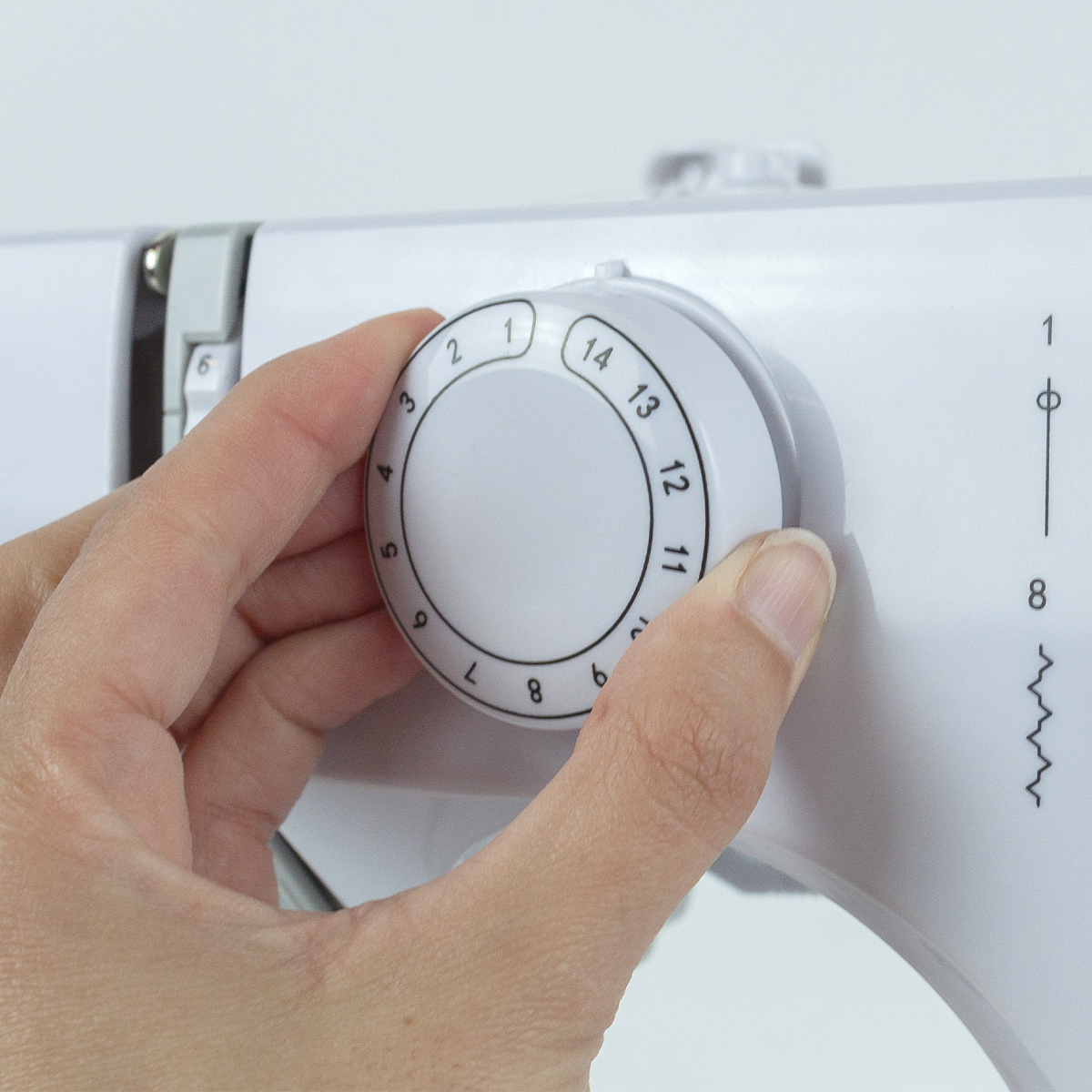 Ręka obraca białe pokrętło na białej maszynie do szycia, pokazując liczby i ustawienia.