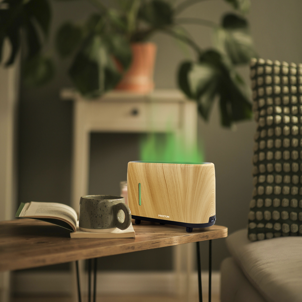 Drewniany dyfuzor zapachów z zielonym światłem, kubek i książka na drewnianym stole. Roślina w tle.