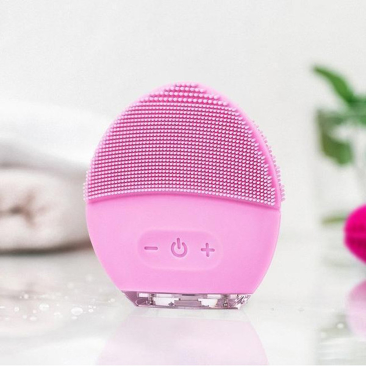 Un cepillo de limpieza facial rosa, ovalado, con cerdas suaves y botones de control. Está sobre una superficie blanca.