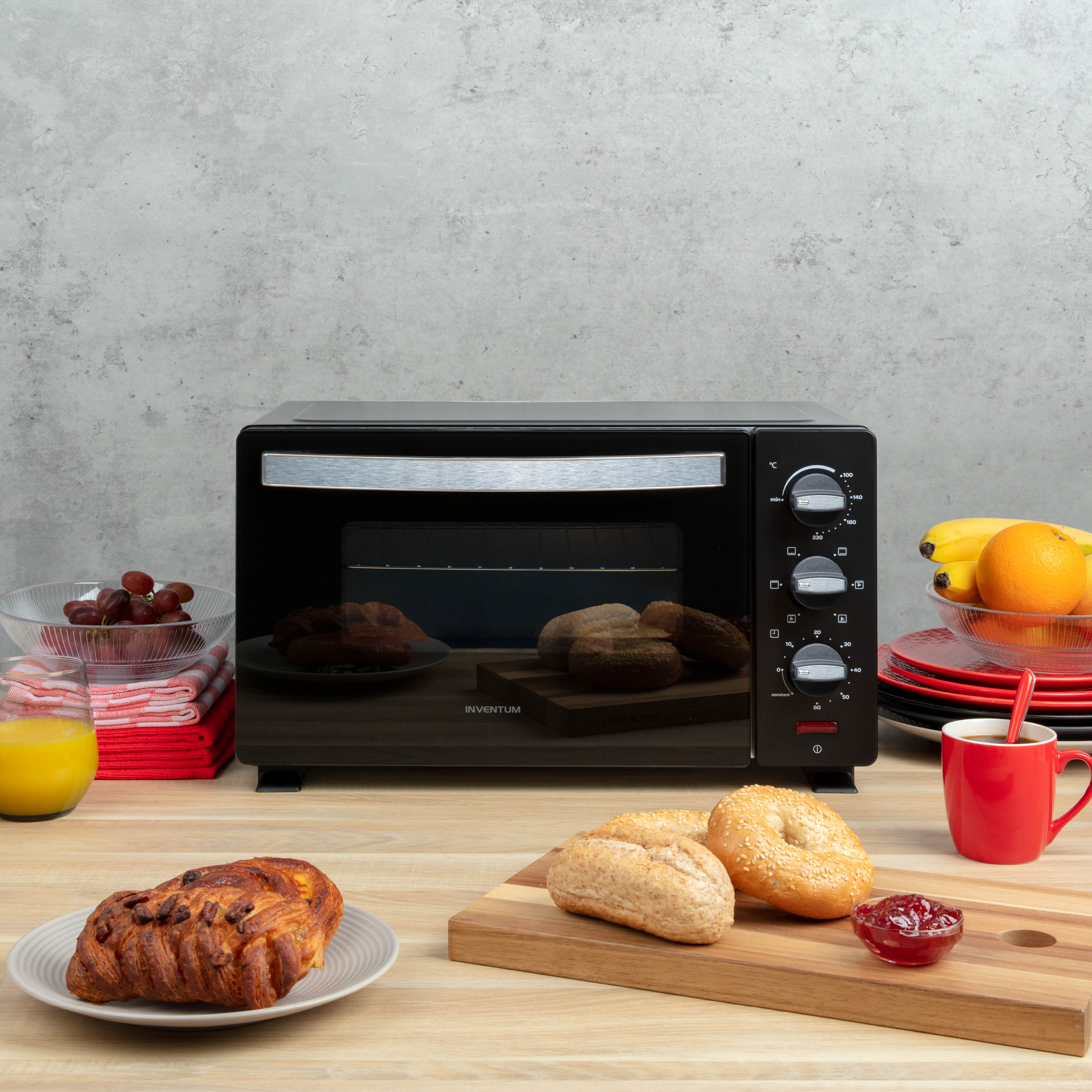 A black toaster oven on a wooden table with food items like bread, pastries, and fruits around it.