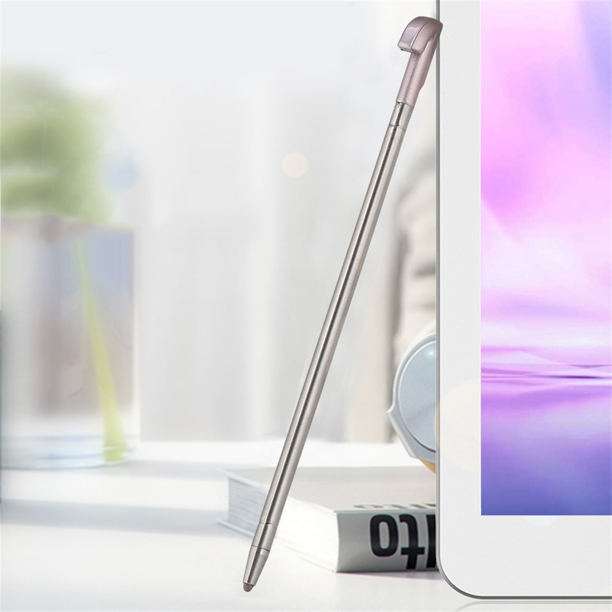Silberner Stylus-Stift lehnt an einem weißen Tablet, auf einem Schreibtisch mit Buch im Hintergrund.