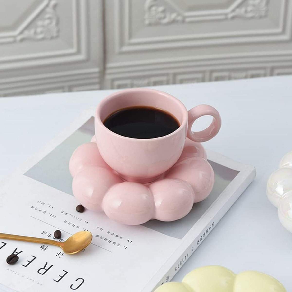 Ein rosa blütenförmiger Untersetzer mit einer rosa Tasse mit dunkler Flüssigkeit auf einem Buch mit goldenem Löffel.