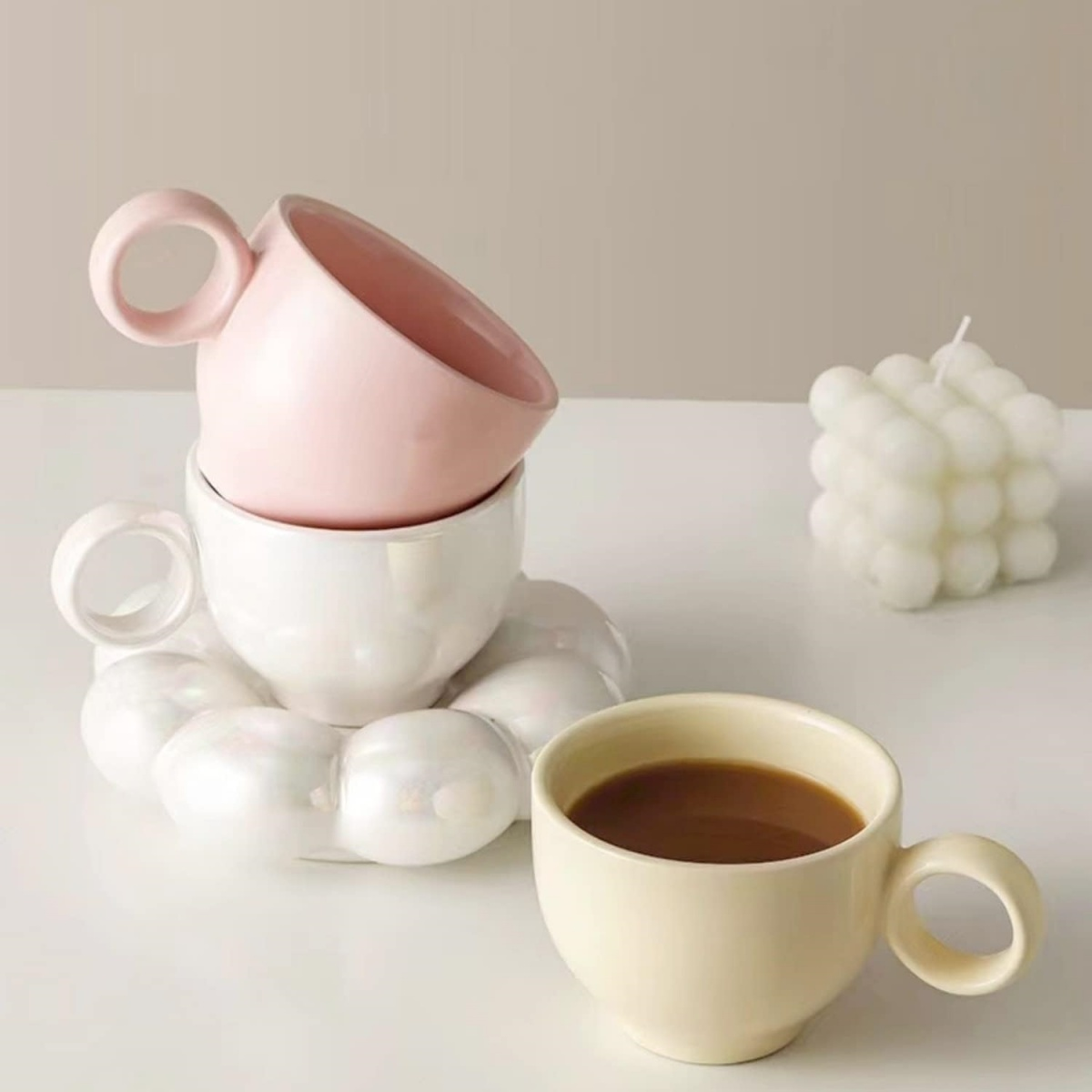 Drei Keramiktassen, eine rosa, eine weiße, eine cremefarbene, stehen auf weißem Untergrund mit einer Kerze.