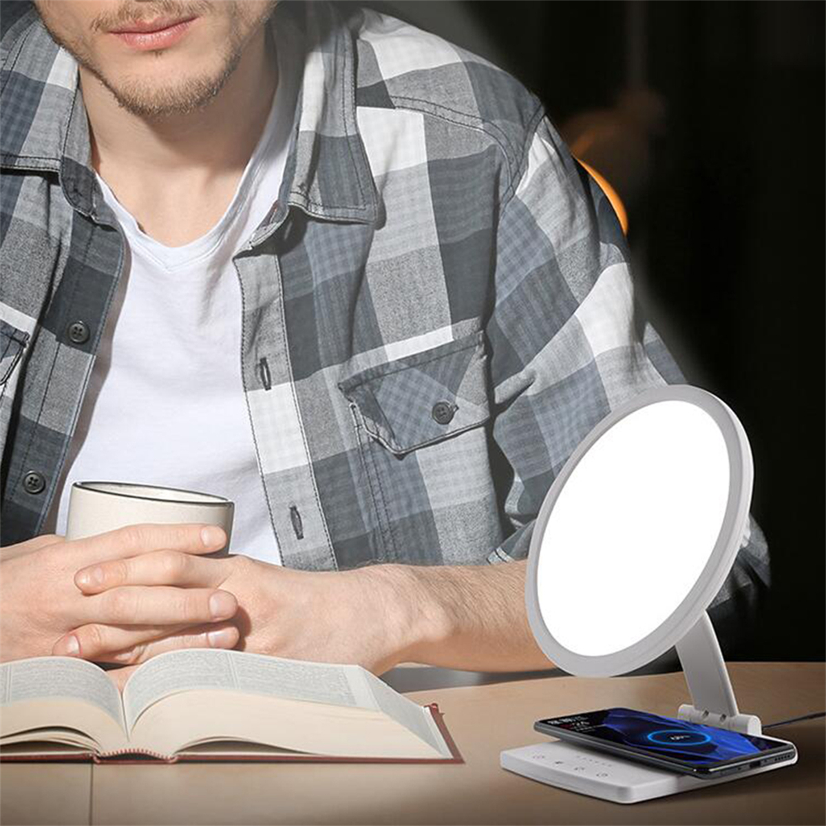 Ein Mann in einem karierten Hemd sitzt an einem Schreibtisch mit einem Buch, einer Lampe und einem Telefon, das auf einem kabellosen Pad geladen wird.