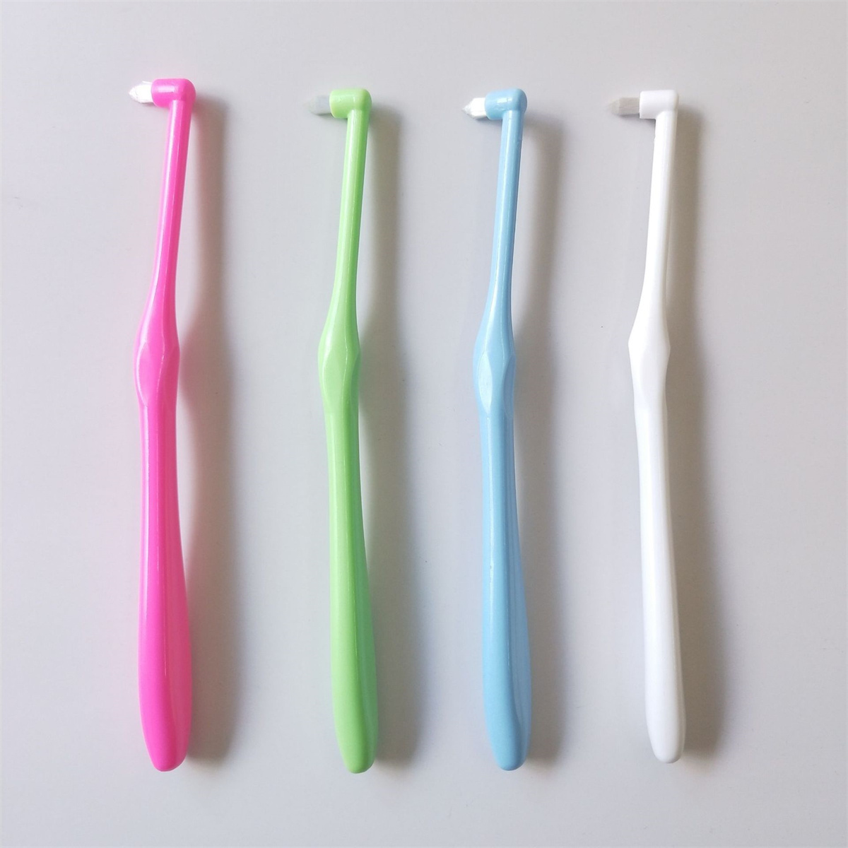 Vier Interdentalbürsten in Pink, Grün, Blau und Weiß, angeordnet auf einer weißen Oberfläche. Jede hat einen Griff.