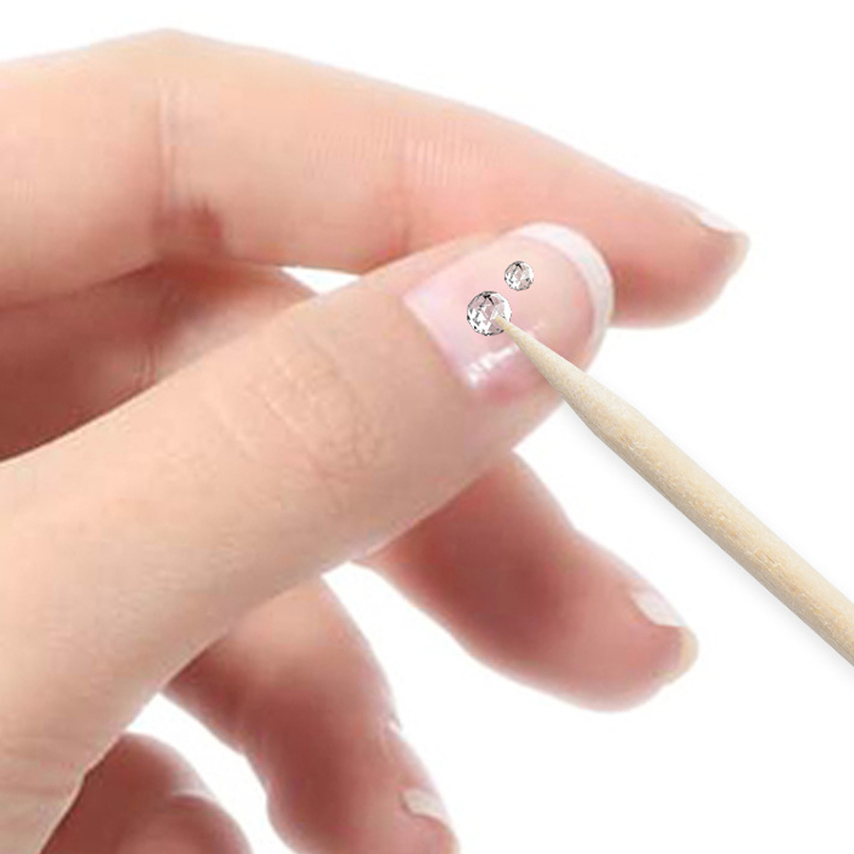 Eine Hand, die einen Nagel mit zwei kleinen Strasssteinen hält, und ein Holzstab neben den Nägeln.