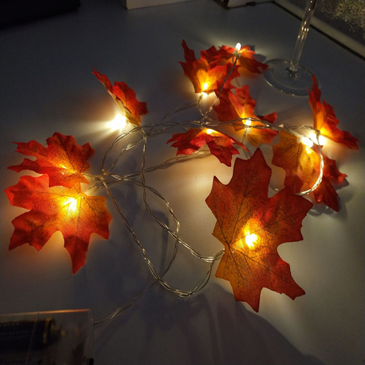 Schnur aus orangefarbenen Ahornblattlichtern auf dunkler Oberfläche. Ein Weinglas befindet sich oben rechts.