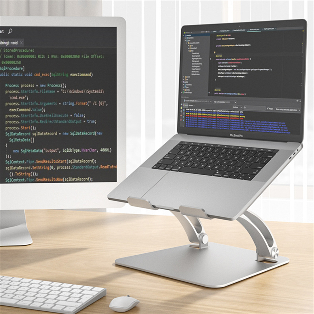 Ein Laptop auf einem silbernen Ständer, mit Code auf dem Bildschirm, neben einem Monitor und Tastatur.