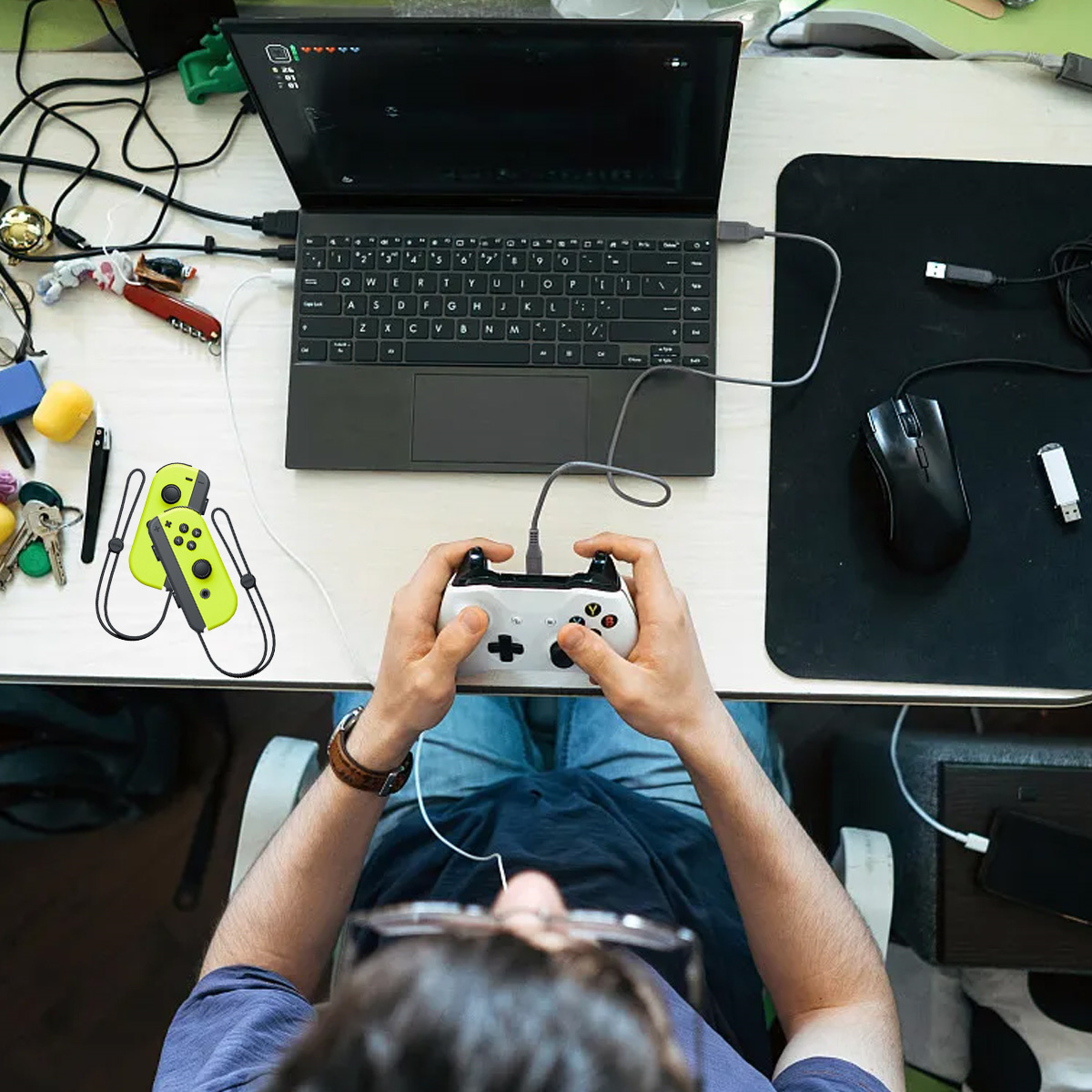 Eine Person mit Brille, die einen Laptop und einen Controller benutzt, mit einem zweiten Controller und Zubehör auf dem Schreibtisch.