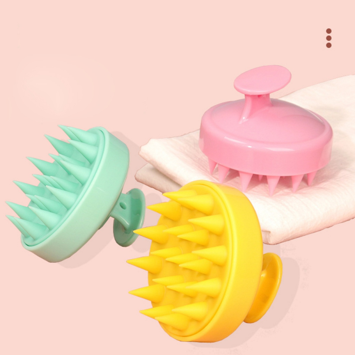 Drei Kopfhautmassagegeräte, grün, gelb und rosa. Die Bürsten liegen auf einem weißen Tuch über einer rosa Oberfläche.