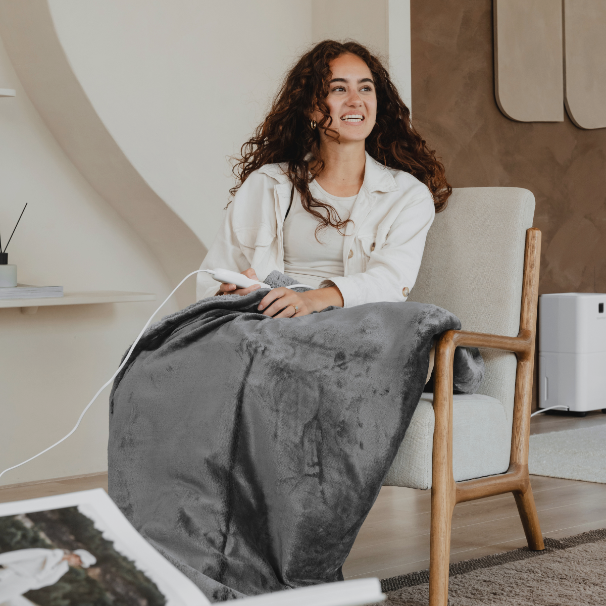 A woman sits, wrapped in a gray blanket, with a remote. A lamp and a book are visible.