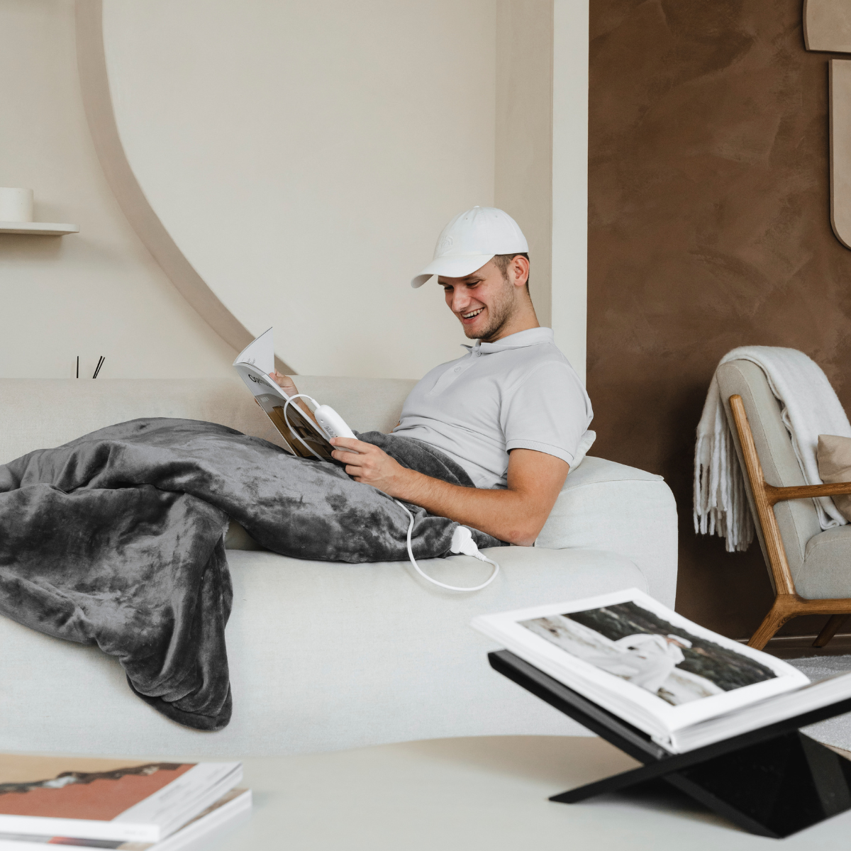 Man on sofa, wrapped in a gray heated blanket, reading a book, wearing a white cap. Book on table.