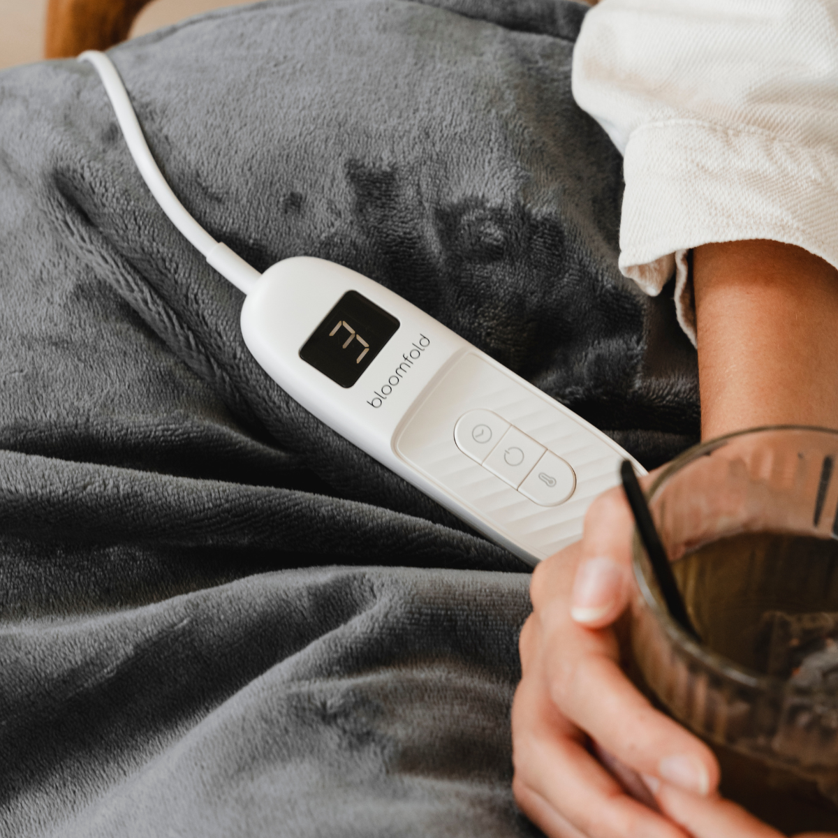 Person holding a glass of tea, with a heating pad controller on a gray blanket.